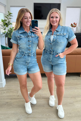 Two women wearing denim rompers taking a mirror selfie in a living room.