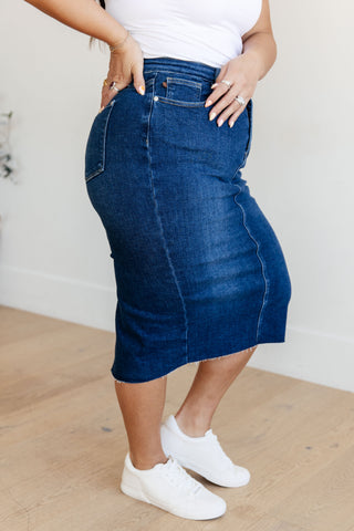 Side view of plus size model wearing a blue denim skirt with white sneakers on a neutral background