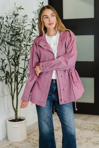 Woman wearing a mauve pink jacket and blue jeans indoors with a plant in the background