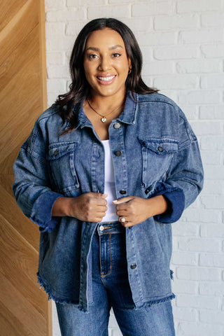 Woman wearing a denim shacket jacket and jeans standing against a white brick wall.