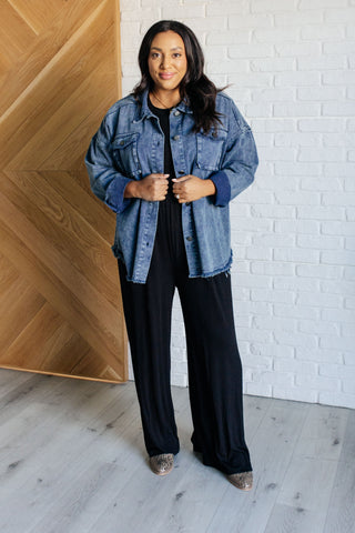 Woman wearing 100% cotton blue denim jacket shacket over a black outfit in a room with wooden and white brick walls.