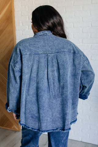 Rear view of person wearing a blue denim jacket shacket with pleat detail in front of a white brick wall.