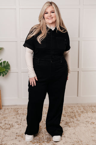 Plus sized woman wearing a black denim jumpsuit with a long white shirt underneath, standing against a light-colored wall.