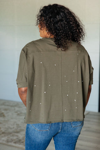 Rear view of person wearing an olive green shirt with white paint splatter design and blue jeans indoors.