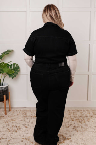 Rear view of plus sized model  wearing a black denim jumpsuit standing in a room with a plant and wooden furniture.