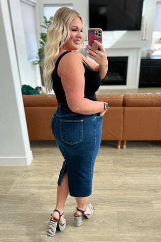 Woman taking a mirror selfie in a living room wearing a black top and blue denim skirt.