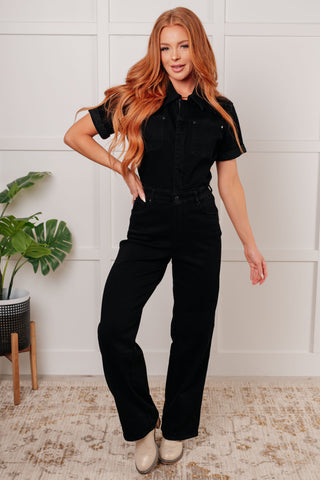 Woman wearing black denim tummy control jumpsuit with short sleeves, standing in a room with a plant and white wall.