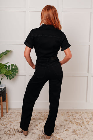 Rear view of model wearing black denim jumpsuit standing in a room with a white wall and plant.