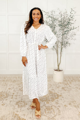 Woman wearing a white dress with maple print, standing in a room with a plant and tiled wall.